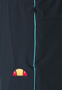 Tmavá tkanina s hladkým povrchem, se tyrkysovým prošíváním a vícolorovou nášivkou (žlutá, červená) poblíž bočního švu.