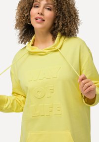 Haut à capuche jaune avec une texture douce, présentant un design en relief avec le texte "CHEMIN DE LA VIE". Comprend une capuche et des cordons de serrage.