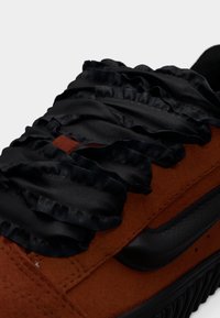 Κοντινό πλάνο καφέ σουέντ αθλητικού παπουτσιού με κορδόνια από μαύρο υφασμάτινο βολάν και μαύρη υφή σόλα, σε λευκό φόντο.