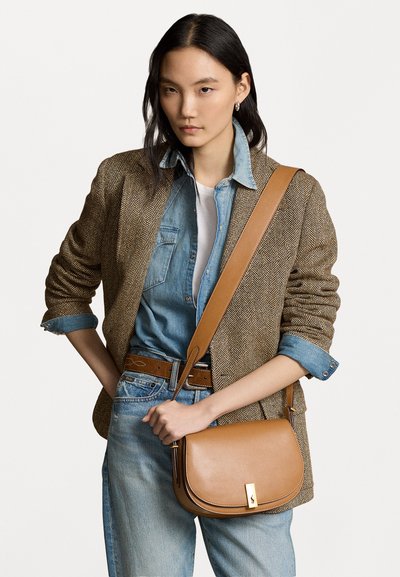 Bolsa de ombro de couro castanho com um formato curvado, detalhes em metal dourado, usada sobre uma camisa de ganga e um blazer em padrão espinha de peixe, combinada com jeans claros.