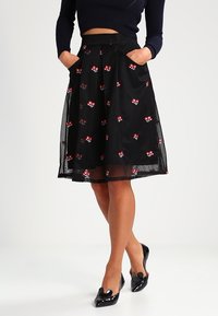 Žena v černé midi sukni s červenou květinovou výšivkou, námořnickém krátkém svetru, se rukama v kapsách a černými lakovanými lodičkami.