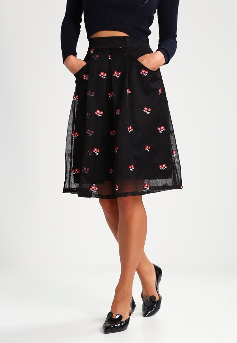 Žena v černé midi sukni s červenou květinovou výšivkou, námořnickém krátkém svetru, se rukama v kapsách a černými lakovanými lodičkami.