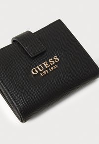 Juodas tekstūruotas piniginė su auksiniu "GUESS EST 1981" logotipu. Yra su užskaitos uždarymu ir kompaktiška stačiakampio forma.