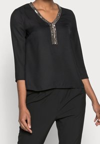 Schwarze Bluse mit V-Ausschnitt, verziert mit silbernen Pailletten entlang der Vorderseite. Dreiviertelärmel, glatte Textur und lockere Passform.