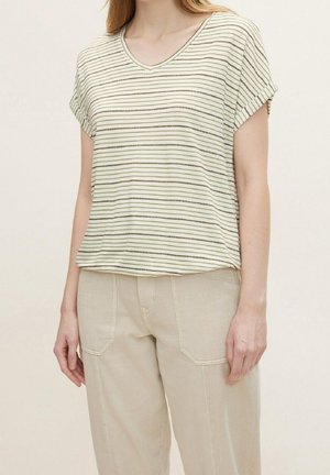 Kobieta ubrana w biało-czarną pasiastą bluzkę z krótkim rękawem i dekoltem w serek oraz beżowe spodnie, stojąca na tle jednolitego jasnego tła.