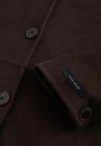 Manche de manteau en laine brun foncé avec des boutons foncés et une étiquette noire portant les mots "FAIT MAIN" cousue près du poignet.