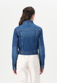 Jaqueta de ganga cropped em azul, com um colarinho clássico, mangas longas e punhos abotoados. A parte de trás apresenta um detalhe em patch centralizado e uma textura suave.