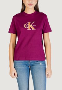 Camiseta de algodón en burdeos intenso con un gran logo dorado "CK" y el texto "Calvin Klein", con mangas cortas y un ajuste relajado.