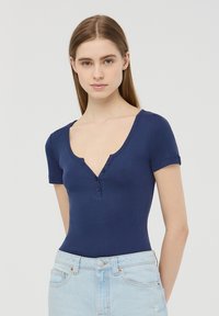Body aderente blu navy con maniche corte, scollatura a V profonda e патн tapo a tre bottoni. Tessuto liscio; abbinato a jeans azzurri a vita alta.