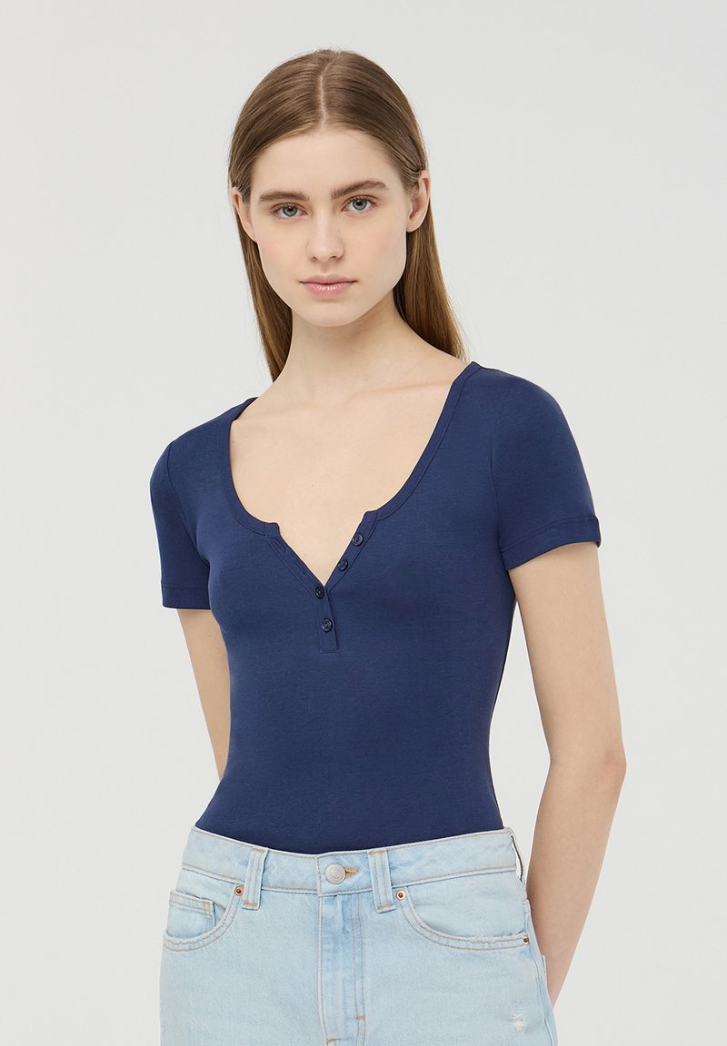 Body aderente blu navy con maniche corte, scollatura a V profonda e патн tapo a tre bottoni. Tessuto liscio; abbinato a jeans azzurri a vita alta.