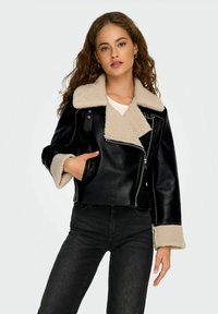 Giacca nera in ecopelle con colletto in shearling beige, design corto, due tasche laterali e chiusura con zip; indossata sopra una camicia bianca.