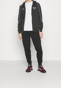 Sudadera con cremallera negra con acentos y logotipos en blanco, combinada con pantalones de chándal negros a juego. Las zapatillas presentan colores negro, rojo y blanco.