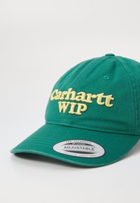 Casquette de baseball en coton vert avec le logo "Carhartt WIP" brodé en jaune, réglable avec une fermeture à pression, visière incurvée et tissu texturé.