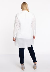 Witte linnen tuniek met lange semi-doorzichtige mouwen, knoopmanchetten en een afgeronde zoom, gecombineerd met donkere spijkerbroeken en zilveren hoge hakken.