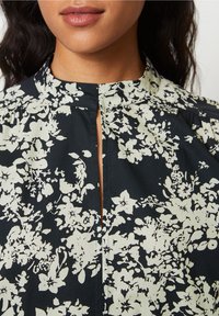 Blouse fleurie noire à fleurs blanches. Dotée d'un col montant et d'une ouverture centrale, avec un tissu doux et texturé ainsi que des détails froncés.