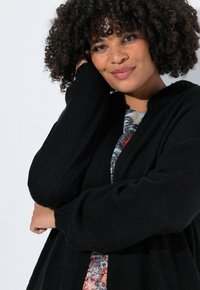 Cardigan noir texturé avec des manches longues, ouvert sur le devant et une coupe confortable, superposé à une robe à motifs avec des accents floraux.
