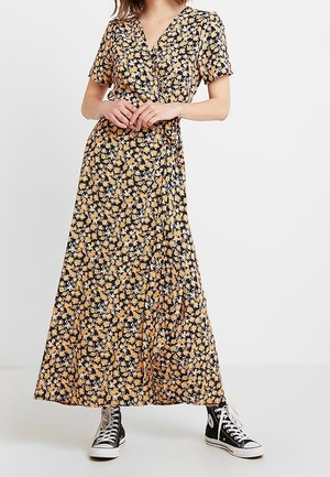 Femme portant une robe portefeuille noire à fleurs avec des manches courtes, associée à des baskets montantes noires, debout devant un fond clair uni.