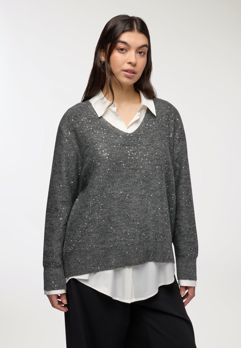 Szary sweter z dekoltem w serek i długimi rękawami, z błyszczącym, teksturowanym wzorem. Nosi się go na białej koszuli z kołnierzykiem, w zestawieniu z czarnymi spodniami.