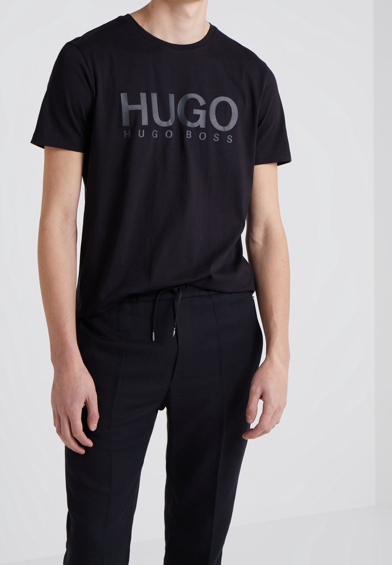 Černé bavlněné tričko s kulatým výstřihem a výrazným tónovým logem "HUGO HUGO BOSS" na přední straně, v kombinaci s černými kalhotami.
