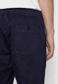 Pantalons bleu marine avec un waistband élastique et une poche arrière à rabat avec bouton. Fabriqués en tissu lisse et léger.