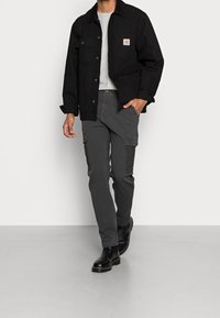 Man som går iförd svart jacka, grå cargo-byxor, ljusgrå skjorta och svarta läderstövlar mot en enfärgad ljus bakgrund.