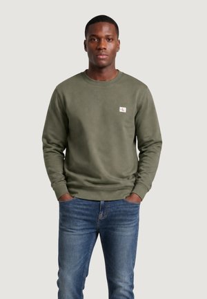 Mand iført en olivengrøn sweatshirt med ck-logo på brystet og blå jeans, stående med hænderne i lommerne mod en ensfarvet baggrund.