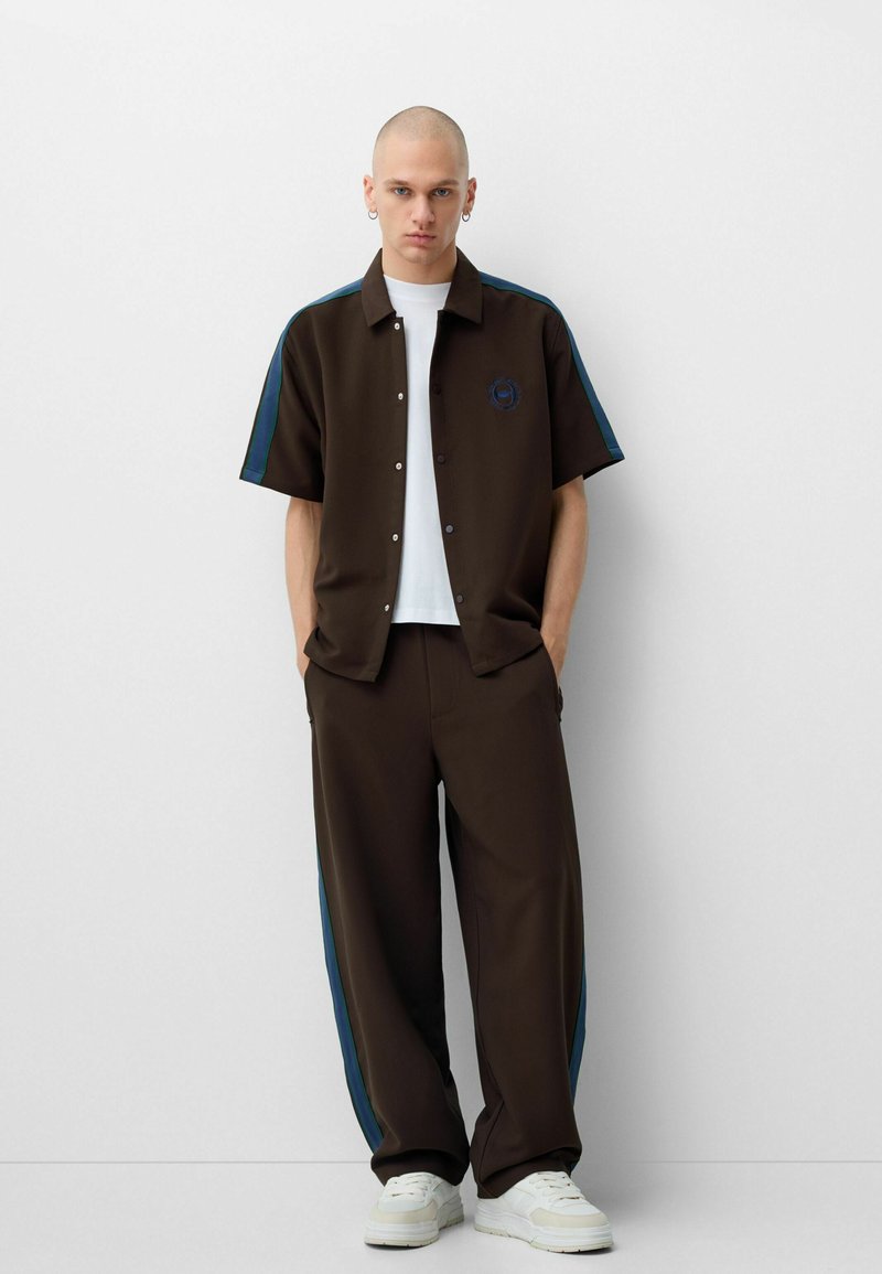 Chemise boutonnée marron avec des accents bleus, associée à un pantalon marron assorti avec des rayures latérales bleues. Un t-shirt blanc et des baskets complètent le look.