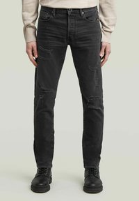 Jeans in denim nero con una vestibilità slim, caratterizzati da dettagli strappati e sbiaditi. Indossati con stivali neri con lacci.