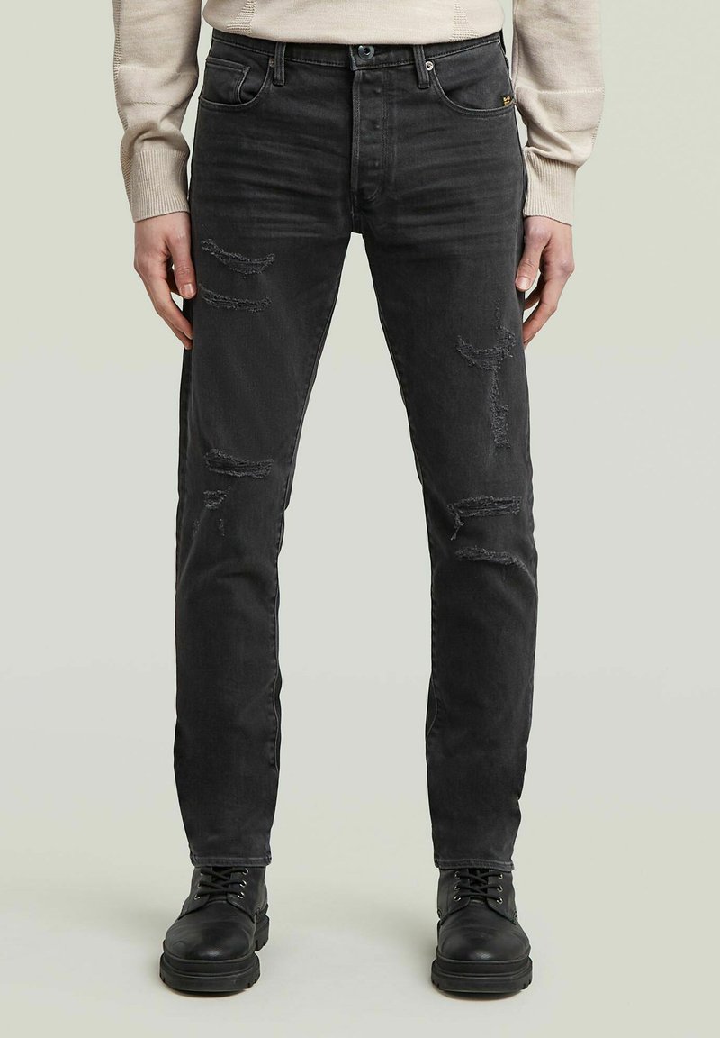 Jeans in denim nero con una vestibilità slim, caratterizzati da dettagli strappati e sbiaditi. Indossati con stivali neri con lacci.
