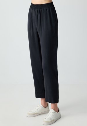 Osoba ubrana w czarne spodnie z prostą nogawką i elastycznym pasem oraz białe trampki, stojąca na tle jednolitego jasnoszarego tła.