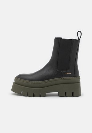 Black leather ankle boots with elastic side panels, thick green rubber platform sole, and pull tab. Features branded detailing on the side.