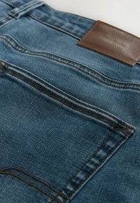 Jeans en denim de couleur bleu foncé avec une finition texturée. Présente une poche arrière surpiquée et une étiquette en cuir marron à la taille.
