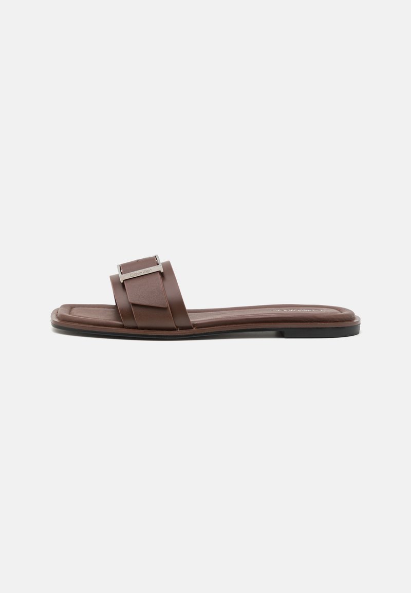 Sandalias deslizantes marrones con dos correas anchas y un acento metálico. La suela es plana, con una textura suave y un diseño minimalista.