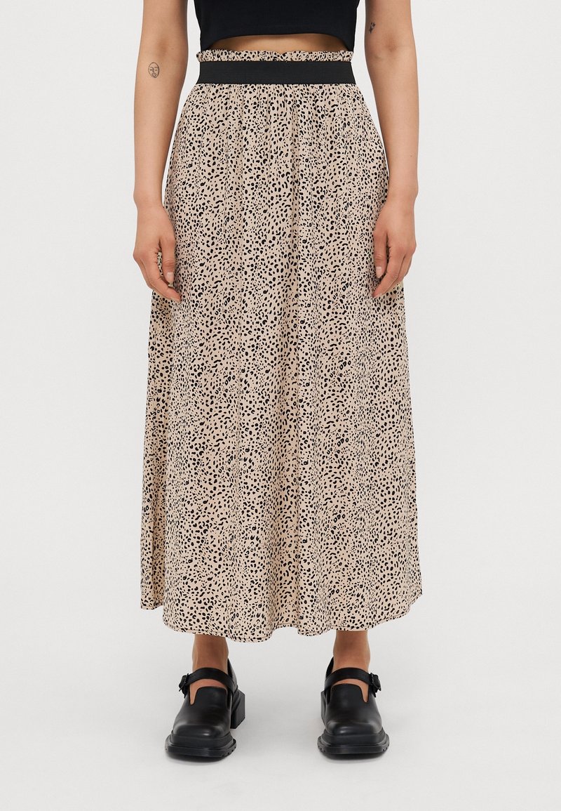 Femme portant une jupe midi fluide beige taille haute à pois léopard noirs, un top court noir et des chaussures noires à boucle, debout sur fond blanc.