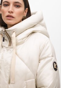 Puffig krämfärgad kappa med en stor huva, guld-färgad dragkedja, beige dragsnoddar och en logotypdetalj på ärmen. Slät, glansig textur.