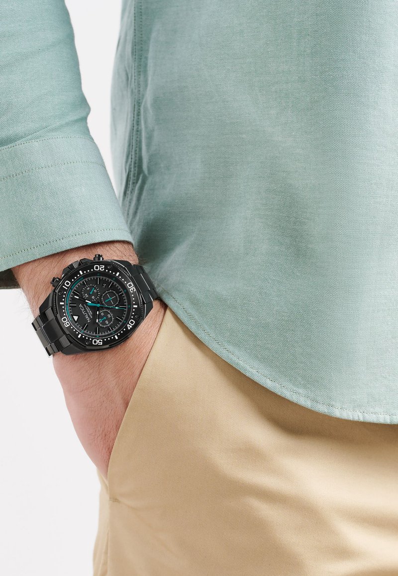 Orologio cronografo nero con accenti turchesi, indossato su un polso, accanto a una camicia verde menta e pantaloni beige. Texture del tessuto liscia visibile.