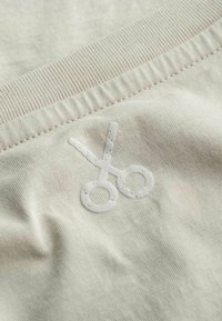 Lichtbeige stof met een zachte textuur, voorzien van een geborduurd ontwerp van een witte schaar en ribbelaccenten langs de rand.