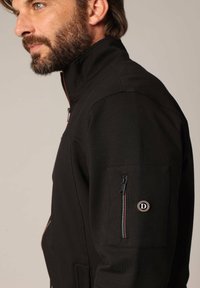 Zwarte ritjack met hoge kraag, een gladde textuur en een zijzak met rits, voorzien van een cirkelvormig logo-detail op de mouw.