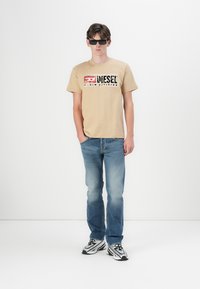 T-shirt en coton beige avec un graphisme noir et rouge "Diesel Denim Division", assorti à un jeans en denim bleu clair et des baskets noir et blanc.