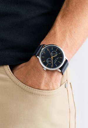 Montre analogique avec un cadran bleu foncé, un boîtier en argent et un bracelet en cuir bleu marine. Comprend deux sous-cadrans temporels et des accents dorés.