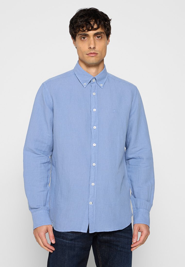 Joven vestido con una camisa de manga larga de botones azul claro y jeans oscuros, de pie contra un fondo claro y liso.