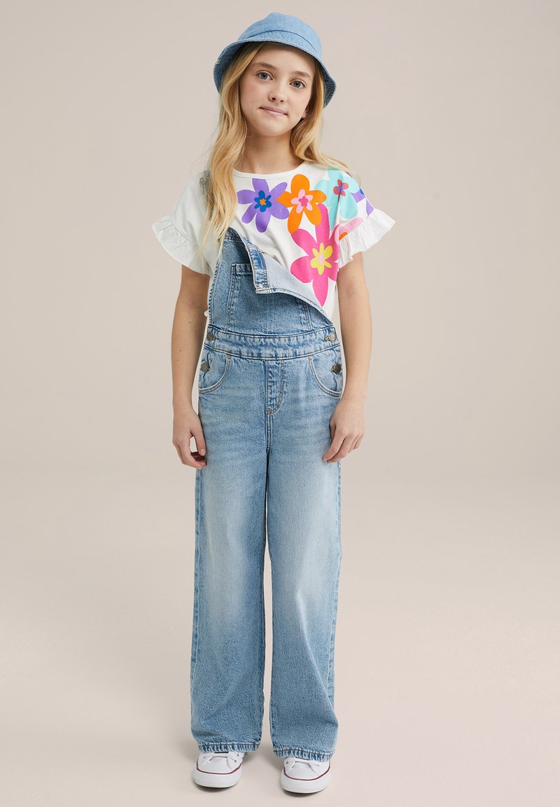 Lichtblauwe denim salopette met een losse pasvorm, gecombineerd met een witte crop top met kleurrijke bloemmotieven. Een lichtblauw vissershoedje maakt de outfit compleet.