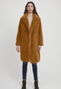 Brun falsk pelsfrakke med en stor krave, dobbeltknaplukning og sidelommer. Båret over en lys sweater og parret med mørke jeans.
