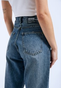 Hoge taille blauwe denimjeans met een vervaagde afwerking, vijf zakken en een zwart merklabel op de achterkant van de tailleband.