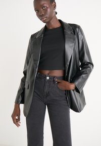 Černý umělý kožený blejzr se strukturovaným designem, s klopami a jednou přední kapsou. Na sobě má černý crop top a tmavé džíny.