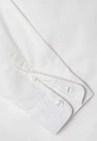 Polsino di camicia bianca con chiusura a bottone, caratterizzato da un tessuto liscio, dettagli cuciti e un bordo arrotondato con una asola.