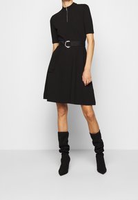 Schwarzes kurzes Kleid mit kurzen Ärmeln, hohem Kragen und Reißverschluss, verfügt über eine taillierte Taille und Taschen. Trägt kniehohe schwarze, lässige Stiefel.