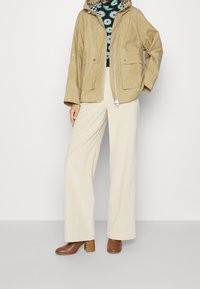 Béžová lehká bunda s kapucí a dvěma předními kapsami, doplněná krémovými širokými kordové kalhotami a hnědými botami na podpatku.