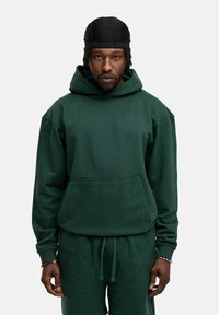 Grüne Fleece-Hoodie mit einer geräumigen Fronttasche und verstellbarem Kordelzug im Kapuzenbereich, kombiniert mit passenden grünen Jogginghosen. Glatte Textur.