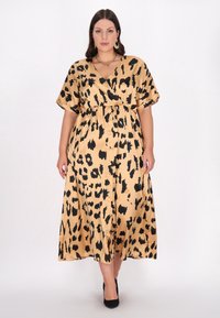 Beige leopardmønstret maxikjole med indpakningsdesign, korte ærmer, en indsnøret talje og en glat, skinnende tekstur. Sorte højhælede sko bemærket.